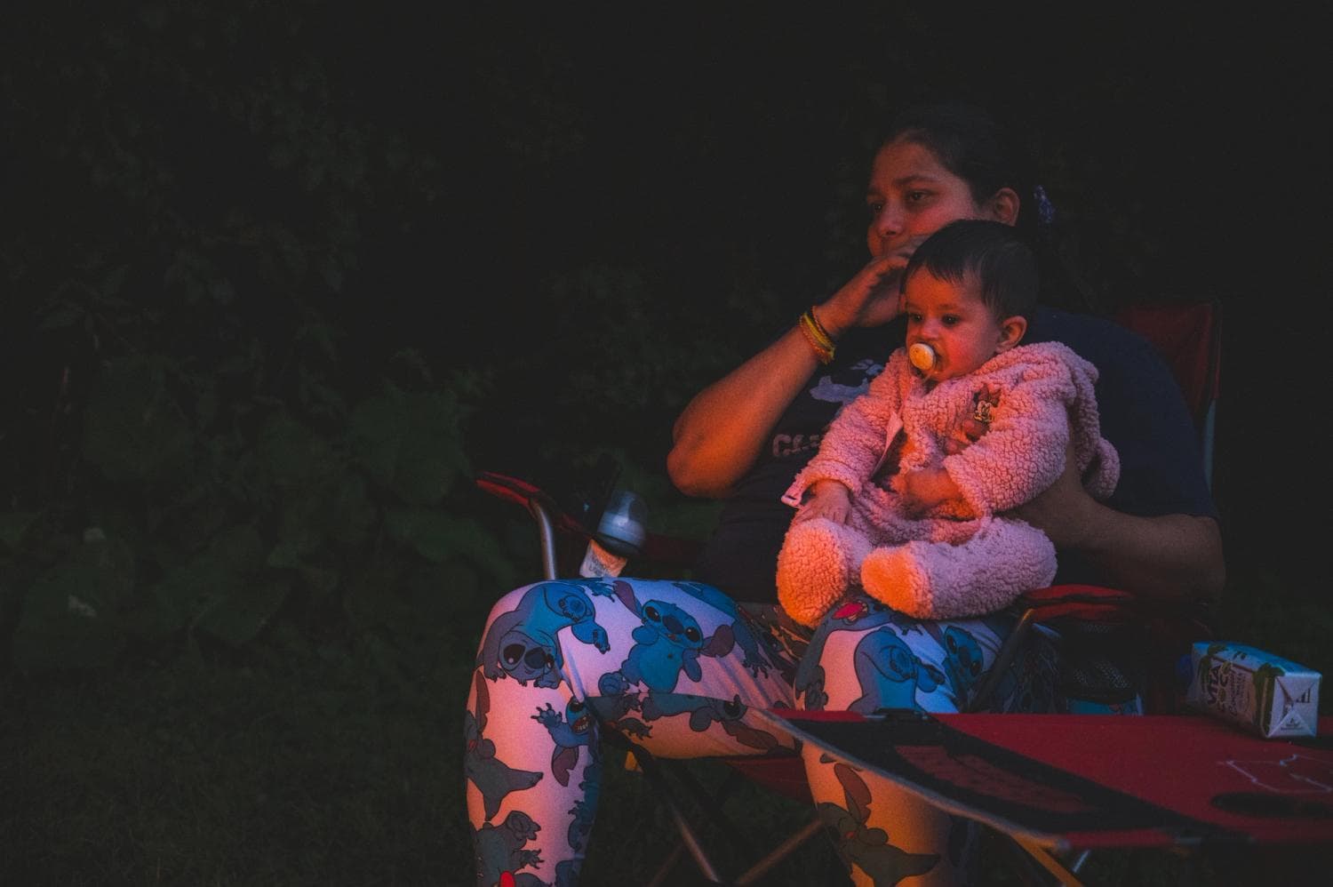 Suveera sitting on her mothers lap in a camping chair starring at a campfire off screen